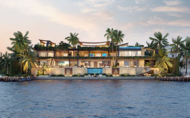 A wide architectural rendering of a contemporary multi-level waterfront residence viewed across calm water. The building has a long horizontal composition, extensive glazing, warm timber-toned finishes, stone or masonry walls, flat rooflines with sculptural canopy elements, balconies, and lush palm landscaping integrated around the facade.