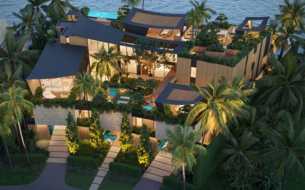 Aerial architectural rendering of a lush tropical coastal resort or multi-residence development. Several interconnected modern buildings feature dark pitched and low-slope roofs, warm timber-clad walls, extensive roof terraces and planted green roofs. Landscaped courtyards, palms, illuminated pathways, pools, and outdoor seating are arranged throughout the property, with the ocean visible beyond.