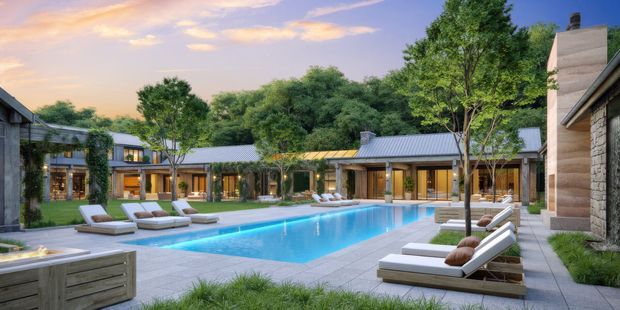 A landscaped courtyard-style outdoor pool area at dusk, surrounded by a low-rise contemporary building arranged around the pool. The scene includes a long rectangular swimming pool, pale stone or concrete paving, multiple cushioned sun loungers, built-in planting beds, mature trees, and warm interior lighting visible through extensive glazing. The buildings feature simple horizontal forms, grey metal pitched roofs, timber-toned framed openings, and textured stone or concrete wall elements.