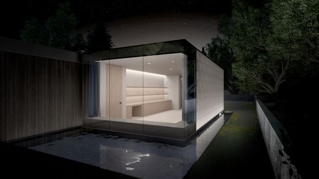 Night-time view of a compact contemporary pavilion or extension with a flat roof and full-height glazed corner. The illuminated minimalist interior appears to contain built-in cabinetry or shelving and a small seating/work area. Dark vertical cladding, pale textured side cladding, reflective paving or water-like surface, and a narrow landscaped path frame the structure.
