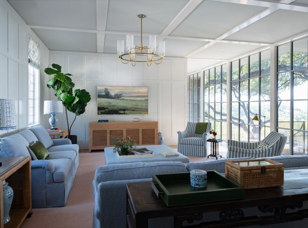 Bright, refined living room with white paneled walls, a coffered ceiling, expansive black-framed multi-pane glazing, and a large seating arrangement around a central ottoman. The room includes blue-grey upholstered sofas and striped armchairs, a rattan-front sideboard, dark timber console table, indoor greenery, landscape artwork, and a brass-and-glass chandelier. The glazing provides abundant natural light and views of trees and water or distant landscape.
