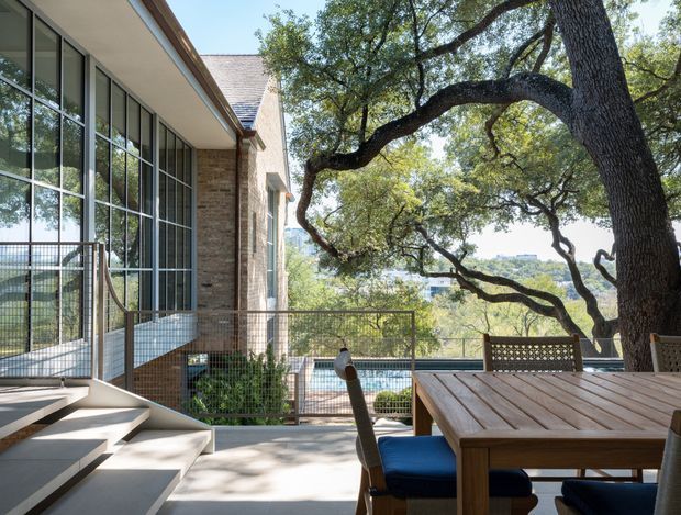 A contemporary multi-level outdoor dining terrace beside a glazed brick-clad building. A large established tree frames the view, while timber decking, a timber dining table, metal balustrades, broad concrete steps, and expansive floor-to-ceiling windows create a bright connection between the building and the landscaped setting. A swimming pool and distant landscape are visible beyond the terrace.