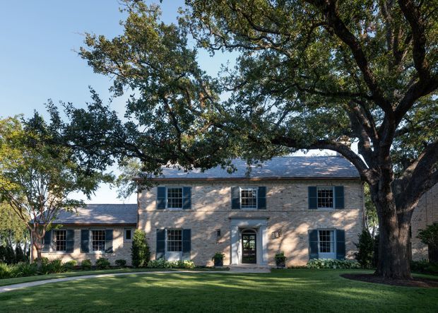 A stately two-storey stone-clad residence viewed from the front, with a symmetrical facade, dark blue-green shutters, multi-pane windows, a central pedimented entry, and a low connecting wing. Mature trees, manicured lawn, and landscaped garden beds frame the home.