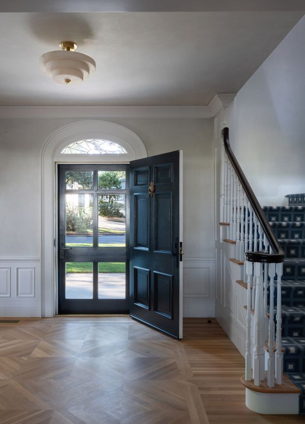 Elegant residential entry hall with a black paneled front door and glazed sidelights beneath an arched fanlight. The space features white painted wall panelling and decorative ceiling cornices, a sculptural flush-mount ceiling light, warm timber parquet flooring, and a staircase with white turned balusters, dark timber handrail, and patterned dark carpet runner. Daylight enters through the open front door.