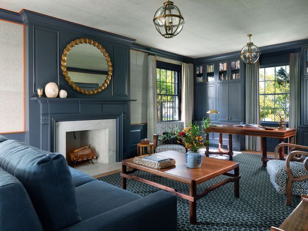 A richly detailed formal living room with deep navy-blue paneled walls, a central fireplace with mantel, tall multi-pane windows, built-in bookcases, and elegant globe pendant lights. A blue upholstered sofa, patterned armchairs, a wooden coffee table, desk, and geometric carpet create a refined, traditional interior with strong symmetry and layered detailing.