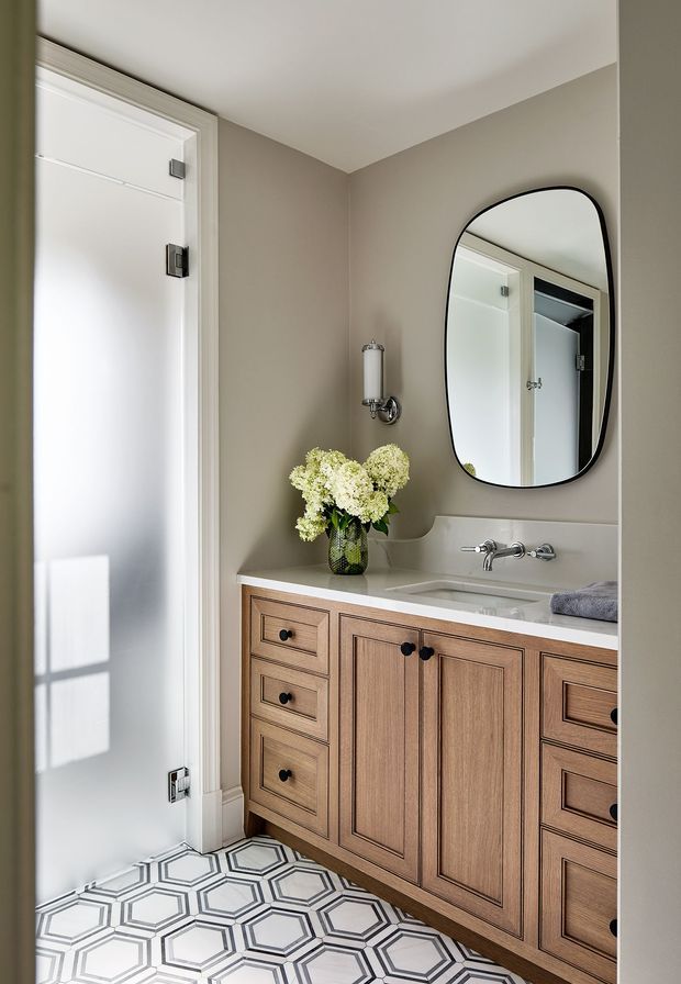 A compact, refined bathroom or powder room with a warm taupe painted wall, white door and trim, patterned geometric floor tile, and a timber vanity with raised-panel drawers and doors. The vanity has a pale stone or solid-surface benchtop, an undermount basin, polished metal tapware, and a rounded rectangular black-framed mirror. A wall sconce and a vase of white flowers complete the understated traditional interior.
