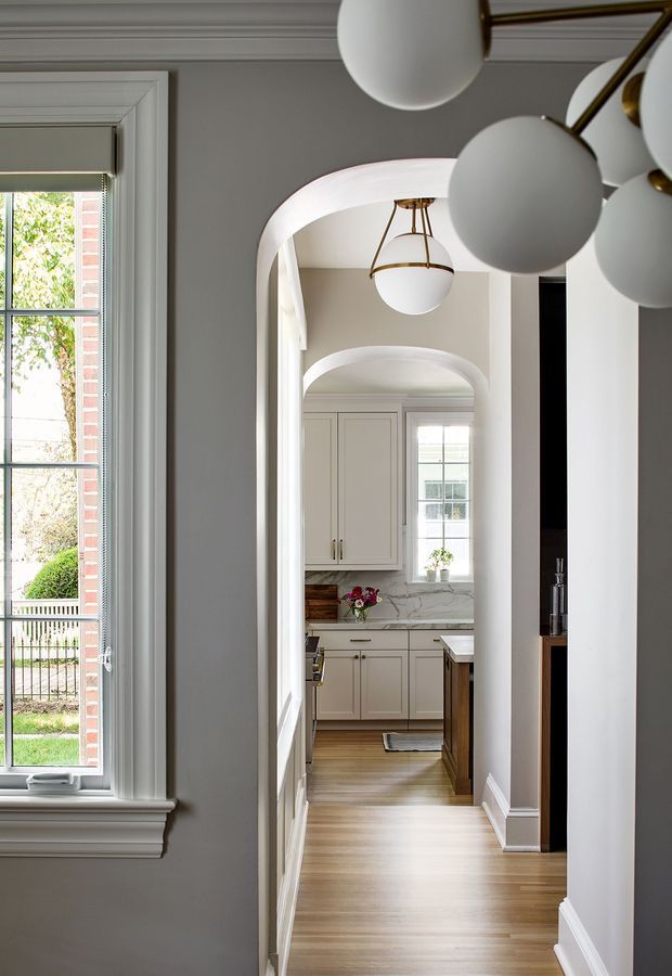 A refined residential interior view through a series of rounded archways, looking from a hallway toward a bright traditional kitchen. The space features grey painted walls, elaborate white trim and mouldings, tall divided-light windows, light timber flooring, cream shaker cabinetry, marble-look benchtops, and elegant globe pendant lighting with brass detailing. The composition emphasises depth, symmetry, and the connection between the hallway and kitchen.