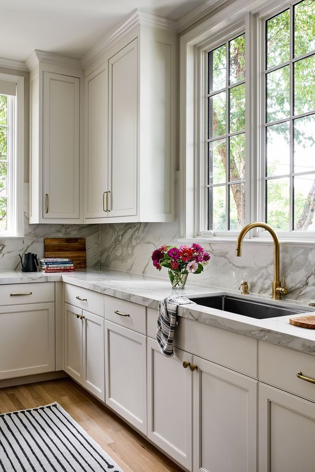 A bright, refined traditional kitchen with cream-painted shaker cabinetry, extensive marble-look stone benchtops and splashback, a deep black sink, and a brass gooseneck tap beneath large black-framed divided windows. Classic panel mouldings, crown cornices, brass hardware, timber flooring, and a striped runner create a polished, welcoming interior. A small flower arrangement and everyday kitchen items add warmth.