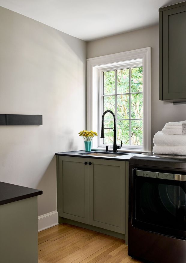 A compact, traditionally detailed kitchen corner with muted sage-green shaker cabinetry, dark benchtops, a black range cooker, and a large white-trimmed divided-light window overlooking greenery. A tall black gooseneck tap sits at the sink beneath the window, with folded white towels stacked on the appliance. The room has warm timber flooring, soft neutral walls, and simple classic detailing.