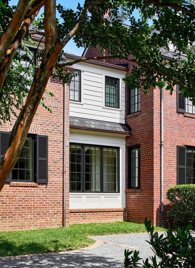 Exterior view of a refined red-brick multi-storey residence, partially framed by mature trees and landscaping. The facade features black multi-pane steel-look windows with shutters, white horizontal siding and trim, a dark shingle-clad projection roof, and a curved gravel walkway leading through the lawn.