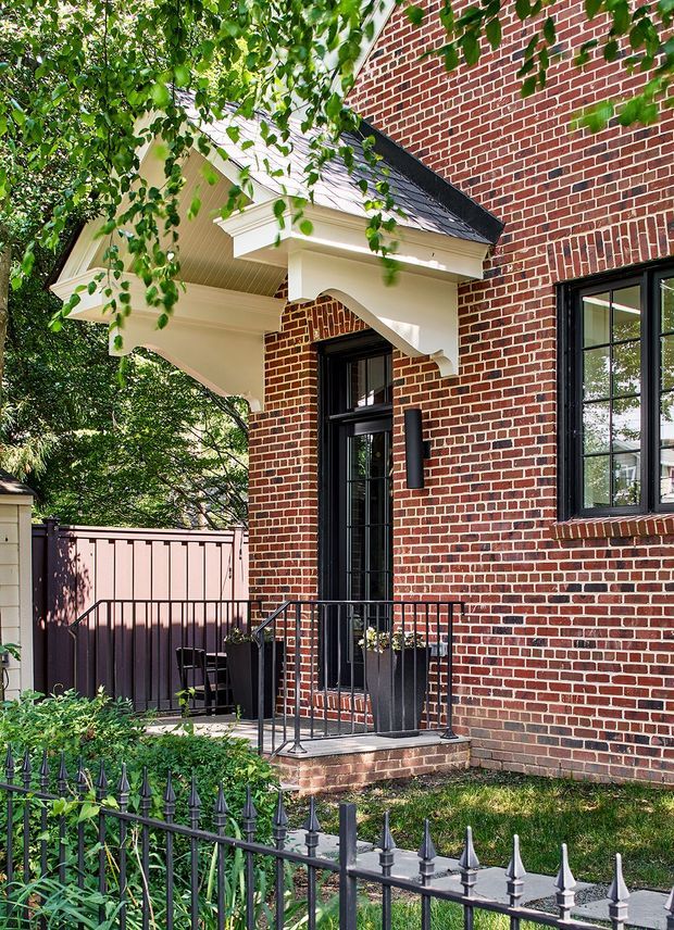 Exterior view of a traditional red-brick residential building entrance, featuring a small covered porch with decorative white timber brackets, a steep dark roof, black-paned windows and door, metal balustrade, brick steps, and a fenced garden courtyard with mature greenery.