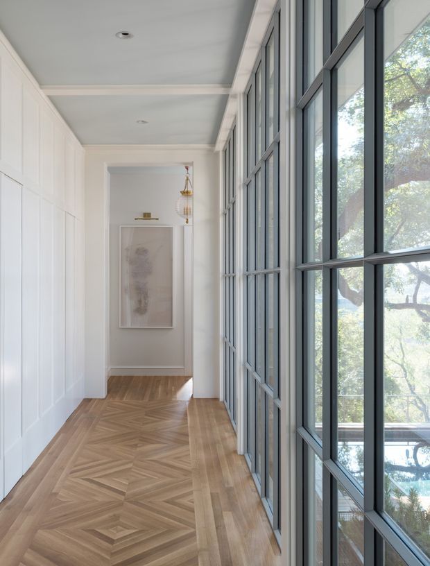 A bright, refined interior hallway with a long wall of dark-framed floor-to-ceiling glazing on one side and white decorative wall panelling on the other. The corridor has a warm patterned herringbone timber floor, simple recessed ceiling lights, a framed artwork, and a delicate pendant light at the far end. Trees and part of an exterior pool or terrace are visible beyond the windows.