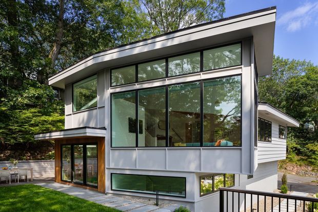 Exterior view of a contemporary multi-level residence set among mature trees. The house features a sharply angled low-pitched roof, extensive floor-to-ceiling glazing, pale horizontal wall cladding, dark trim, and a timber-framed glazed entry projecting from the lower level. A paved walkway, lawn, and balcony railing are visible in the foreground.