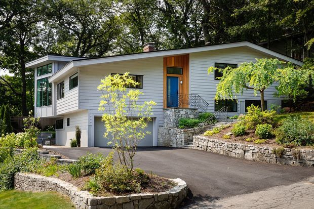 Exterior view of a contemporary single-storey residence set among mature trees on a sloped site. The home has white horizontal cladding, a low-pitched roof with broad eaves, expansive corner glazing, a blue entry door with timber surround, and an integrated single garage. Stone retaining walls, planted garden beds, steps, and a paved driveway define the landscaped approach.