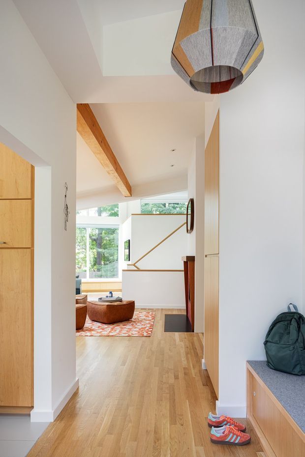 Bright contemporary residential hallway and entry viewed toward an open living area. The space features wide light timber flooring, white walls and ceiling, built-in light timber cabinetry and bench storage, exposed timber ceiling beam, large windows, and a distinctive geometric pendant light. A staircase with a simple timber handrail is visible beyond, along with a seating area and patterned rug.