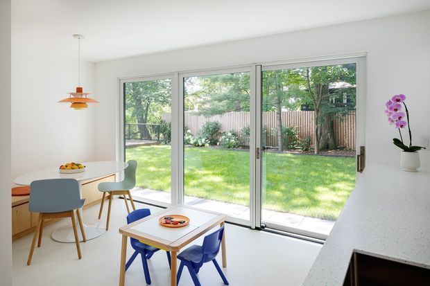Bright contemporary kitchen and dining area opening directly onto a landscaped backyard through large sliding glass doors. The room has white walls and cabinetry or benchtops, pale flooring, light timber dining furniture, colourful chairs, and a distinctive orange pendant light. A white stone-look benchtop occupies the right foreground, with an orchid adding a small decorative accent.