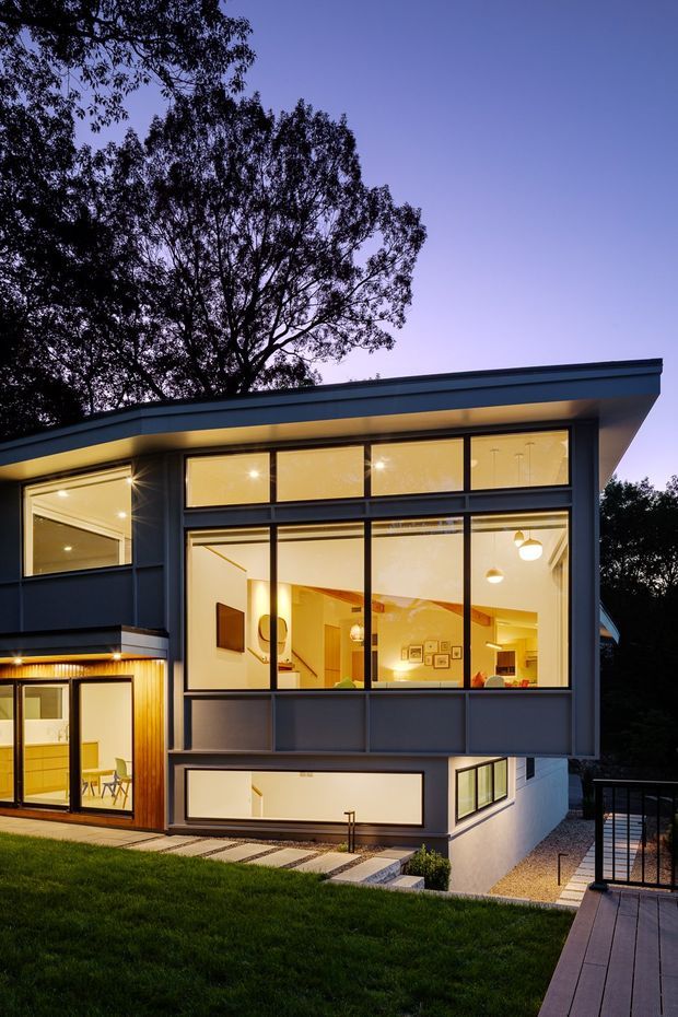 Twilight exterior view of a contemporary two-storey residence set among mature trees. The home has a low-pitched roof with broad eaves, dark grey-blue cladding and framing, extensive warm-lit glazing, a timber-clad entry section, and a raised deck with dark metal balustrading. Interior living spaces are visible through the large windows, with landscaped lawn and gravel surrounding the house.