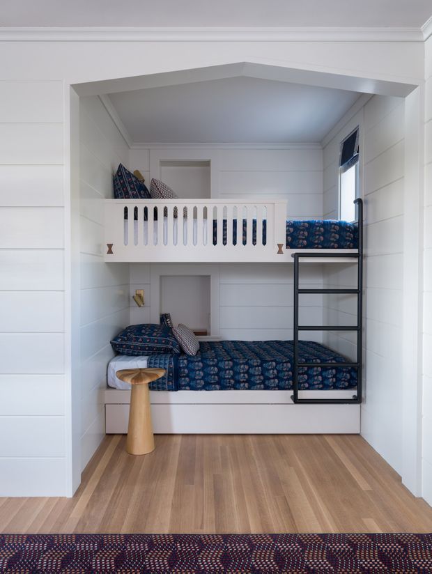A compact bunk bedroom with two built-in sleeping levels, white vertical wall panelling, a white guardrail on the upper bunk, and a black metal ladder. Both bunks are dressed in dark blue patterned bedding, with a small wooden stool beside the lower bed. The room has warm timber flooring, white trim, and a simple, practical built-in design.