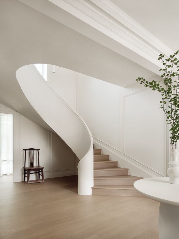 A bright, refined interior hallway and stairway dominated by a sculptural curved white staircase with a smooth balustrade and cylindrical newel. Pale timber flooring, white panelled walls, decorative ceiling moulding, and a dark traditional chair create a restrained classic setting. A small round white console table and tall indoor plant sit at the right edge.