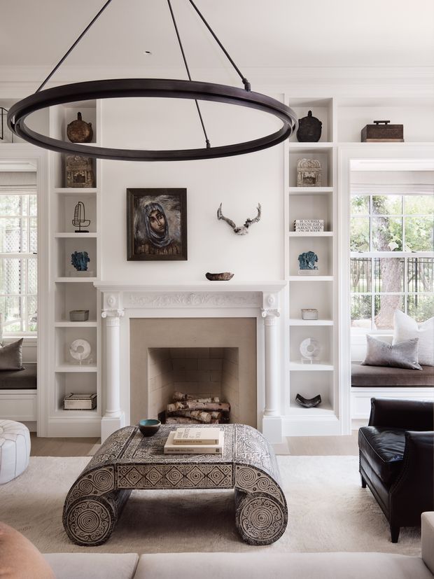 Bright, refined living room centered on a white decorative fireplace with built-in open shelving and window seats on either side. A large dark metal circular pendant hangs above an ornate carved wooden ottoman or coffee table, with leather and upholstered seating, curated objects, artwork, and antler decor. The room has a predominantly white and neutral palette with dark brown and black accents.