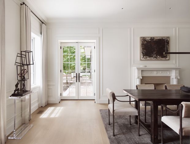 Elegant formal dining room with a dark timber dining table and upholstered chairs, pale timber flooring, white paneled walls, tall curtains, French doors opening to an outdoor area, and a decorative classical fireplace mantel. A sculptural metal artwork and framed artwork add restrained decorative detail.