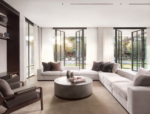 A bright, refined contemporary living room with a large L-shaped upholstered sectional arranged around a round coffee table and a leather-framed lounge chair. Floor-to-ceiling glazed doors and windows open directly onto a landscaped garden, while sheer curtains, dark built-in shelving, recessed ceiling lighting, and neutral soft furnishings create a calm, spacious interior.