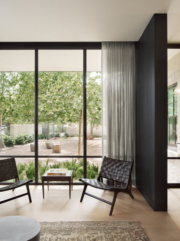 A refined sitting area with two dark woven lounge chairs and a small wooden side table, positioned beside full-height glazed doors or windows overlooking a landscaped courtyard. Pale flooring, sheer pleated curtains, dark window frames, and a prominent dark timber-clad structural column create a calm, modern interior with strong indoor-outdoor connection.