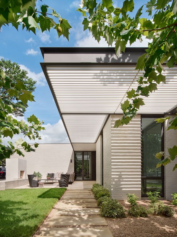 Exterior view of a contemporary single-storey residence framed by leafy trees, showing a broad flat roof with a slatted pergola or covered outdoor walkway. Light textured masonry walls, dark window frames, a paved path, manicured lawn, and a small outdoor seating area define the approach to the house.