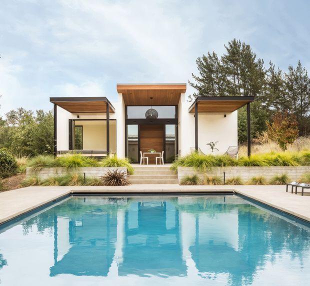 Exterior view of a low, modern pavilion-style home or pool house behind a rectangular swimming pool. The building has white rendered walls, broad flat projecting roof canopies with exposed timber soffits, dark slender structural posts and frames, and a central glazed entry with timber cladding. Layered steps, pale paving, ornamental grasses, and mature planting connect the house to the pool terrace.