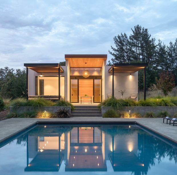 A contemporary single-storey house viewed from across a swimming pool at dusk. The symmetrical facade features a central glazed entry volume with warm timber-toned cladding and lighting, flanked by projecting flat-roofed wings with covered outdoor areas. Concrete steps and raised landscaped garden beds lead down to the rectangular pool, which reflects the illuminated building.