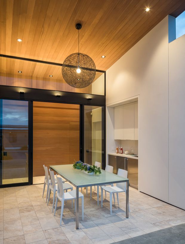 A contemporary dining area with a long glass-topped dining table and white chairs, set beneath a large woven spherical pendant light. The space features a warm timber-lined ceiling, pale stone or porcelain floor tiles, white walls, dark-framed full-height glazing and doors, and an integrated kitchenette or serving nook. A high-level opening brings in additional daylight.