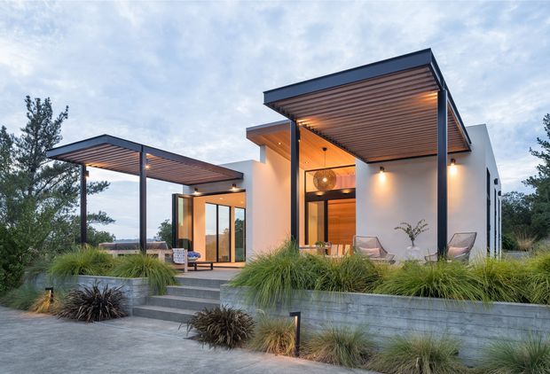 Contemporary single-storey residence with crisp white rendered walls, dark metal framing, and broad flat roof canopies featuring warm timber soffits. Floor-to-ceiling glazing opens onto a stepped concrete entry terrace, while ornamental grasses and low landscaping surround the elevated home. Warm architectural lighting glows beneath the canopies and inside the living spaces at dusk.