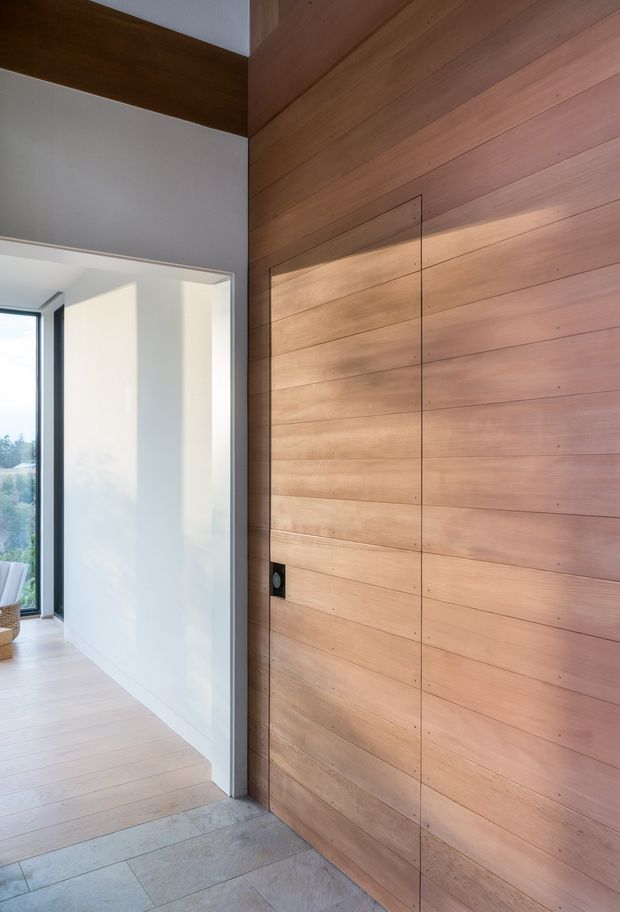 Interior view of a refined entryway featuring a large flush timber pivot door with horizontal wood panelling, set beside a bright white hallway and full-height glazing. Pale timber flooring, grey stone-like entry flooring, slim dark door hardware, and clean linear detailing create a warm, minimalist contemporary feel.