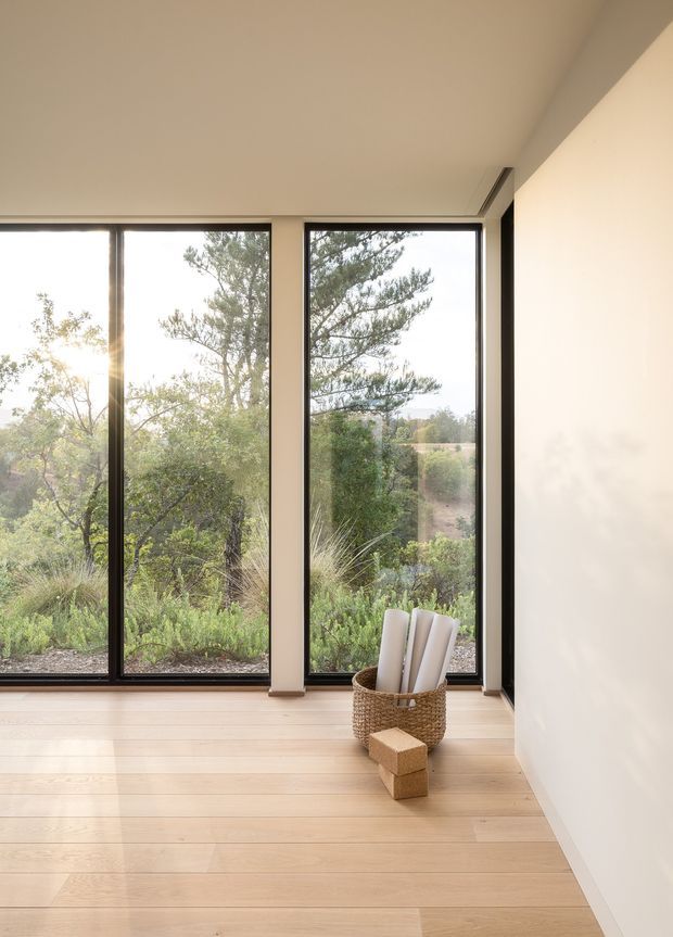A bright, minimalist interior corner with pale timber flooring, smooth warm-white walls and ceiling, and full-height black-framed windows overlooking native landscaped vegetation. A woven basket holding rolled materials and two small wooden blocks sit on the floor, while the simple glazing and clean junctions create a calm connection to the outdoors.