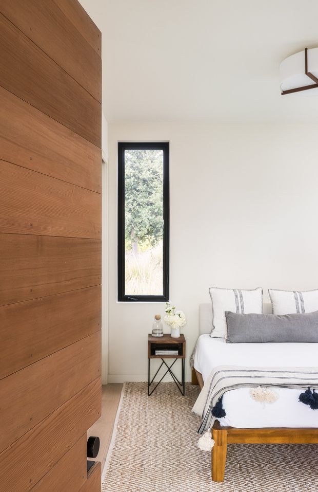 Bright, narrow bedroom with a simple white bed dressed in white and grey striped linens, a small dark wood bedside table, woven neutral rug, and a large built-in or wall-mounted warm timber panelled feature on the left. A slim black-framed window overlooks greenery, while a sculptural ceiling light adds a subtle mid-century touch. The room has a calm, refined, natural feel with clean contemporary detailing.