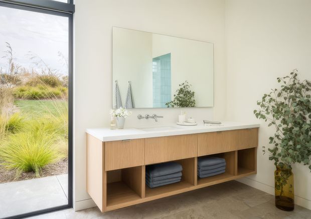 A bright, refined bathroom with a floating double-sink vanity in warm timber cabinetry, open storage cubbies, a white benchtop, wall-mounted chrome tapware, and a large frameless mirror. A full-height glazed door or window opens to a landscaped garden, bringing in abundant natural light. Neutral finishes and neatly folded towels create a clean, calm atmosphere.