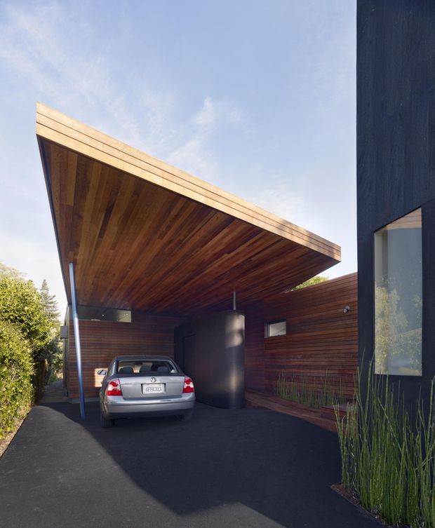 Exterior architectural photograph of a contemporary residence with a dramatic cantilevered timber-lined carport canopy. The dark-stained timber soffit and horizontal timber-clad walls frame a driveway and parked car, while a dark vertical facade with a tall glazed opening appears at right. Dense planting borders the driveway.
