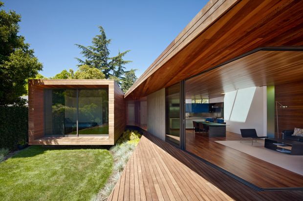 A contemporary single-level residential courtyard viewed from an exterior timber deck. The home features extensive floor-to-ceiling glazing, rich horizontal timber cladding and soffits, a projecting flat roof, and a compact glazed timber-clad pavilion across the landscaped courtyard. The open interior reveals a minimalist living area and kitchen, while the foreground is defined by a broad timber deck and lawn.