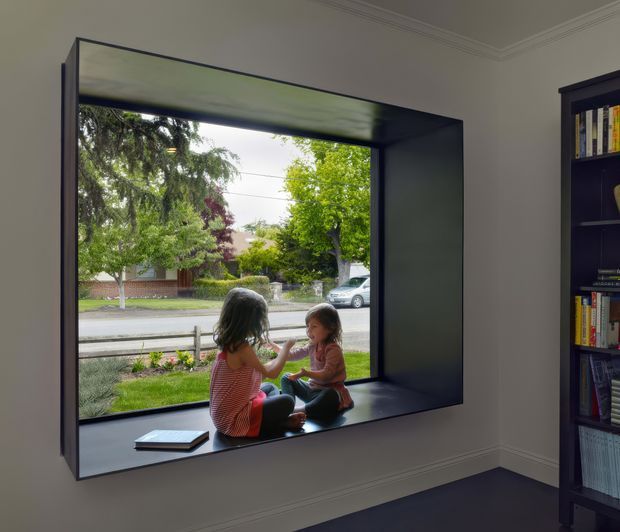 Interior view of a refined residential room featuring a deep, dark-framed recessed window with a broad built-in sill, where two children are seated. The window looks onto a leafy suburban street and landscaped front garden. A tall dark built-in bookcase filled with books occupies the right side, while pale walls, decorative cornice moulding, and dark carpet create a calm, traditional interior.