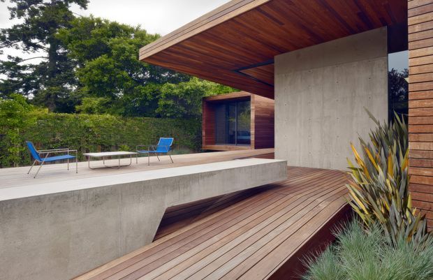 A contemporary flat-roofed home with a dramatic projecting timber-lined soffit, raw concrete walls and terrace, and warm timber decking. The outdoor terrace includes minimalist lounge furniture and overlooks a dense landscaped garden, while large glazed openings connect the exterior to the interior.