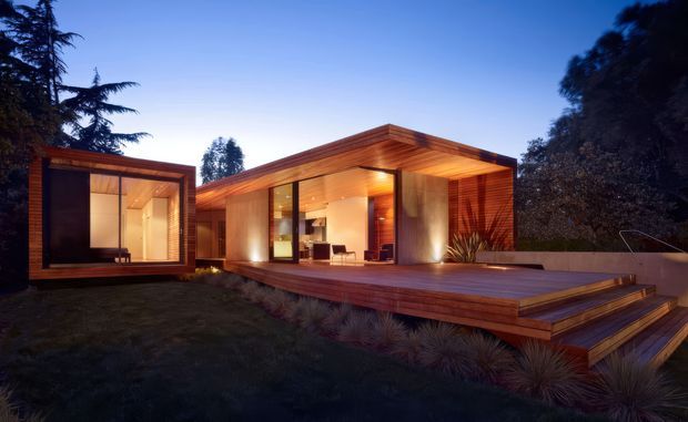Twilight exterior view of a contemporary single-storey house composed of low, rectilinear timber-clad volumes. Large glazed openings reveal a warm, wood-lined interior, while broad timber decking and stepped platforms extend into the landscaped garden. The flat roof and strong indoor-outdoor connection give the home a modern, pavilion-like character.