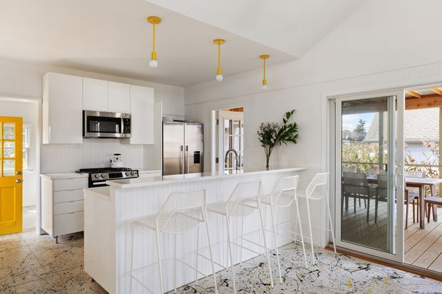 Bright open-plan kitchen with white floor-to-ceiling cabinetry, stainless-steel appliances, a large white island with integrated bar seating, and distinctive yellow pendant lights. The space opens to a timber outdoor deck through glazed sliding doors, with a dining area visible beyond. White vertical wall panelling, patterned flooring, and a light neutral palette create a fresh, airy contemporary interior.