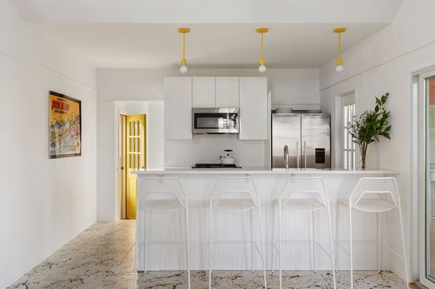 Bright, compact contemporary kitchen with a long white island and four white bar stools. The space features white flat-panel cabinetry, a built-in microwave, stainless-steel refrigerator, pale stone or terrazzo-look flooring, and three pendant lights with yellow accents. A glass-panel door and windows bring in natural light, while a tall indoor plant and framed artwork add warmth.