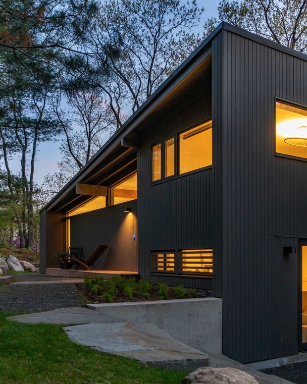 A contemporary dark-clad house viewed from the landscaped exterior at dusk. The building has a low-slope roof, strong rectilinear volumes, vertical dark cladding, warm amber light glowing through windows and recessed outdoor areas, and a concrete path and retaining planter leading through a wooded site. The architecture is understated and modern, with sheltered outdoor seating beneath a deep roof overhang.