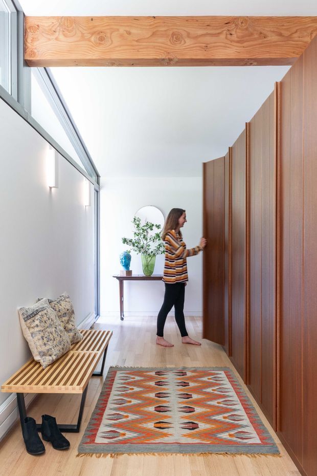 A bright, narrow residential hallway with pale timber flooring, a full-height bank of warm timber built-in wardrobes on the right, and a large glazed opening on the left. A timber ceiling beam spans the corridor, with a slatted timber bench, patterned cushions, shoes, a console table, plant, mirror, and rug adding lived-in detail. A person is opening one of the wardrobe doors.