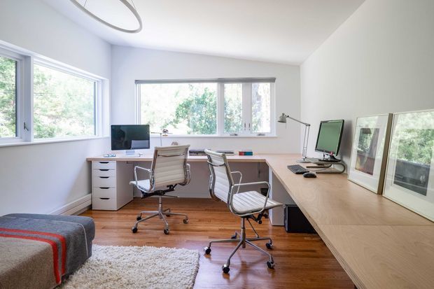 Bright home office with a large L-shaped built-in timber workstation, two computer setups, white drawer units, and two modern swivel office chairs. White walls and expansive windows provide abundant natural light and views of greenery, while polished timber flooring and a textured area rug add warmth. A partial upholstered seating area is visible in the foreground, and framed artwork or prints rest on the desk near the right-side windows.