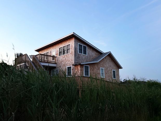Exterior view of a multi-level coastal-style house set amid tall dune grasses. The home has shingle-clad walls, several pitched gabled roof sections, white-trimmed windows, and a raised timber balcony with exterior stairs. Clear blue sky and natural vegetation surround the building.
