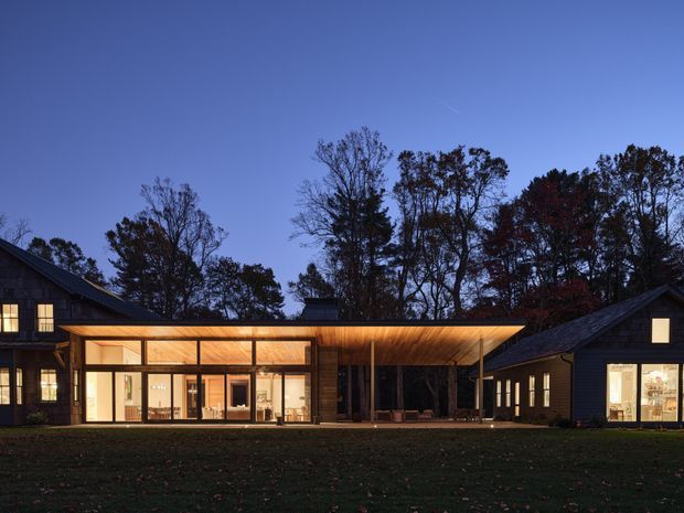 Twilight exterior view of a low, linear residential building complex set among mature trees. Dark timber-clad gabled wings flank a brightly illuminated glazed central pavilion with a broad, shallow projecting roof, creating a contemporary connection between the structures.