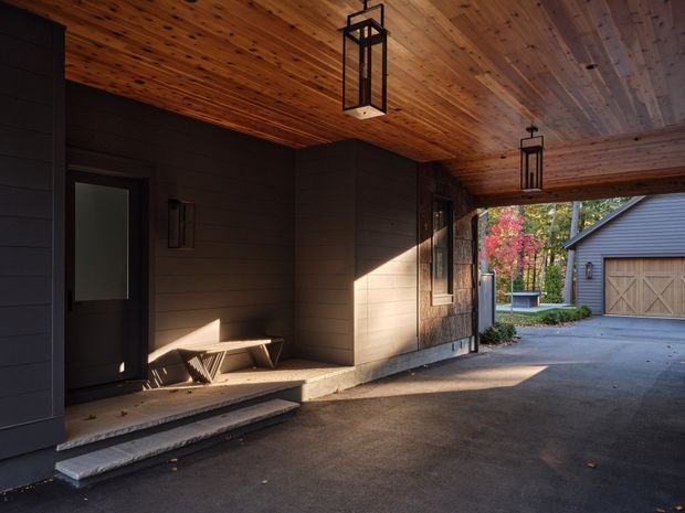 Covered residential entry or carport area beside a dark-clad building, with a warm timber-lined ceiling, hanging lantern-style lights, a small bench, steps, and a driveway leading toward a detached garage. Autumn planting is visible beyond the covered space.