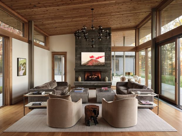 A sophisticated living room with expansive floor-to-ceiling glazing, a warm timber-lined ceiling, and a central dark stone fireplace with a television above it. Two curved upholstered lounge chairs face a pair of sofas around a large rug and low coffee tables. A sculptural clustered pendant light hangs centrally, while views of trees and a garden are visible through the windows.